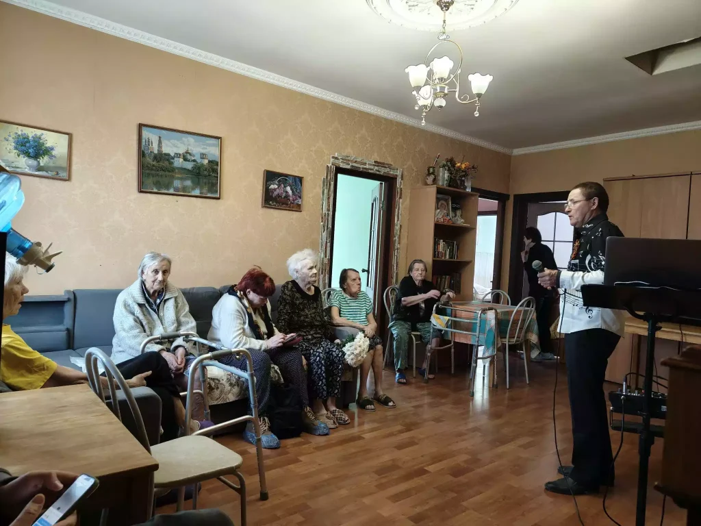 Пожилые люди сидят и слушаю артиста, который стоит перед ними с микрофоном
